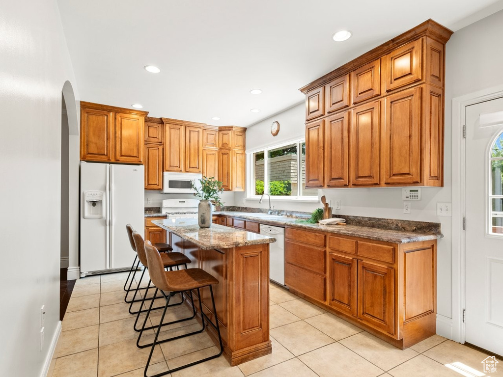 Kitchen featuring white appliances, a center island, a breakfast bar area, brown cabinetry, and light stone countertops