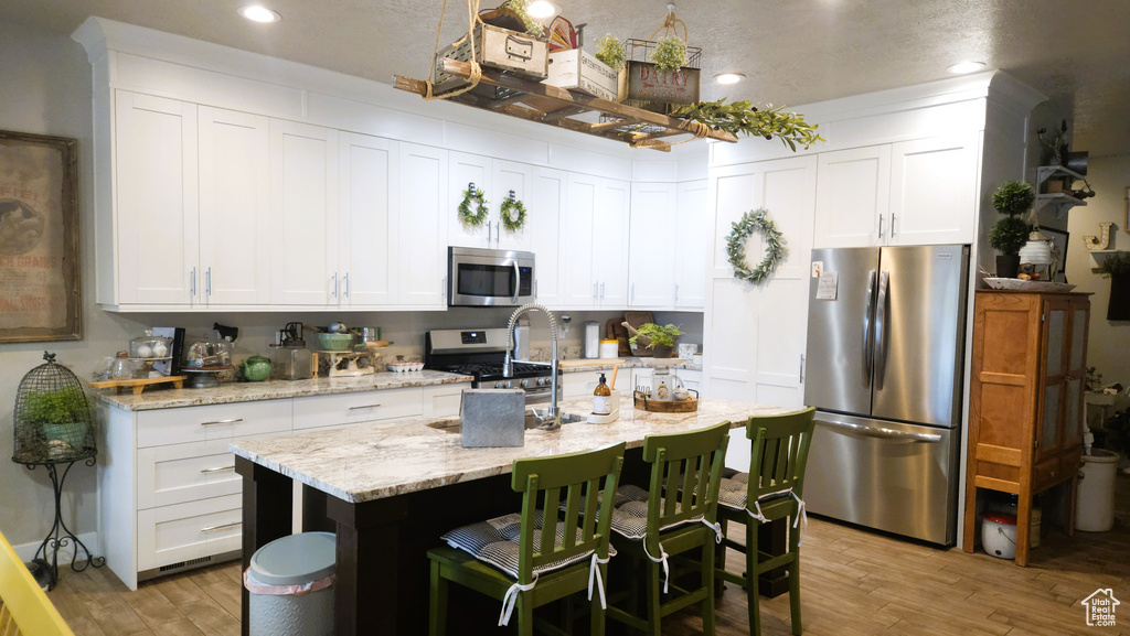 Kitchen featuring appliances with stainless steel finishes, light wood-style floors, a kitchen island with sink, white cabinets, and a kitchen bar