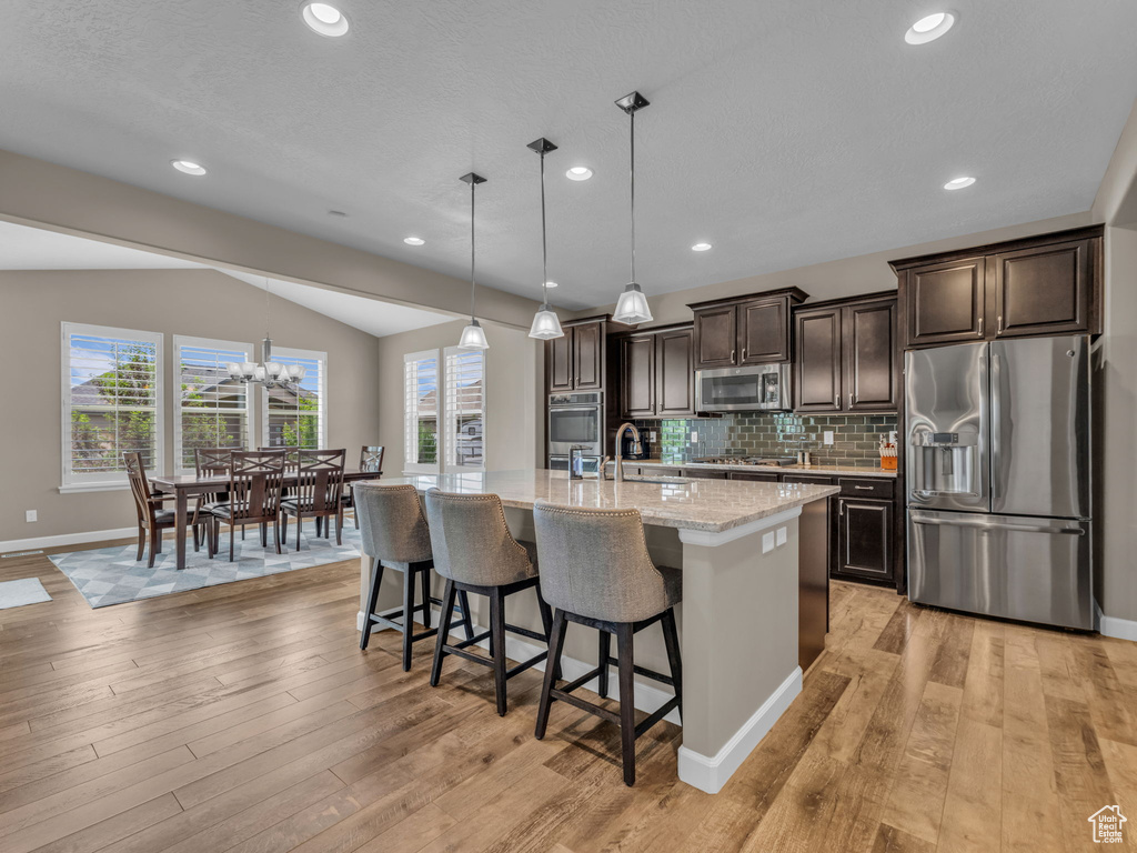 Kitchen featuring stainless steel appliances, dark brown cabinets, lofted ceiling, light wood-style flooring, and a kitchen breakfast bar