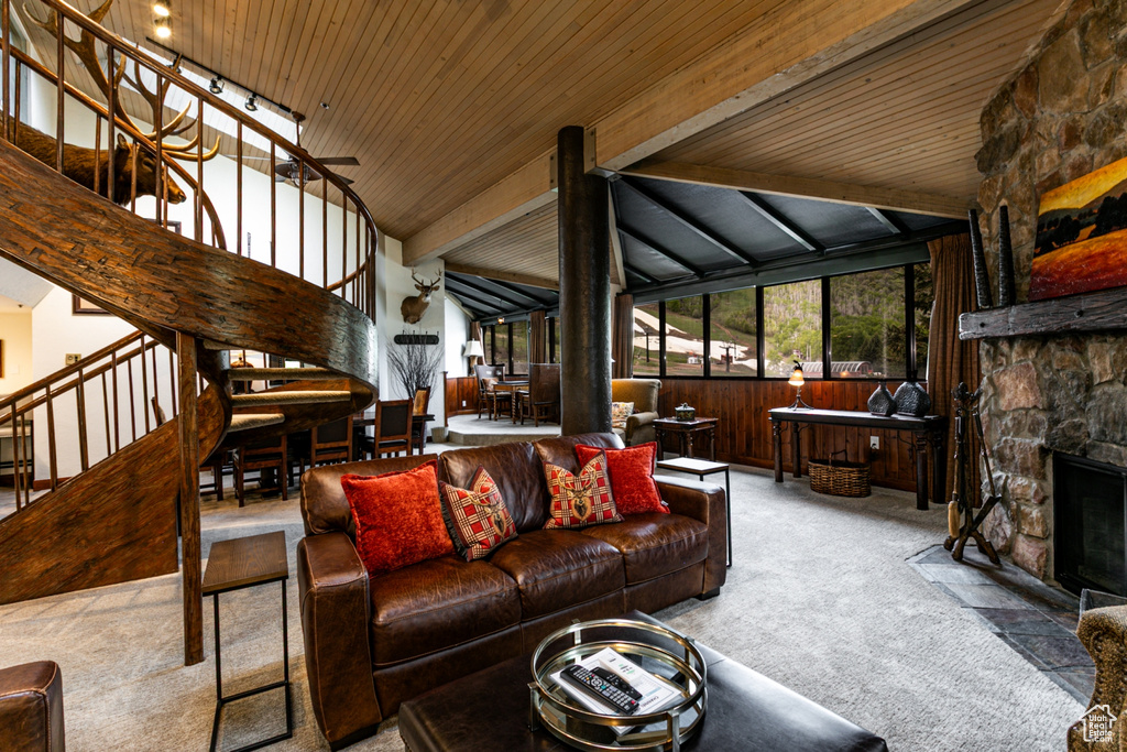 Carpeted living room with stairs, a stone fireplace, wooden ceiling with exposed beams, wood walls, and high vaulted ceiling