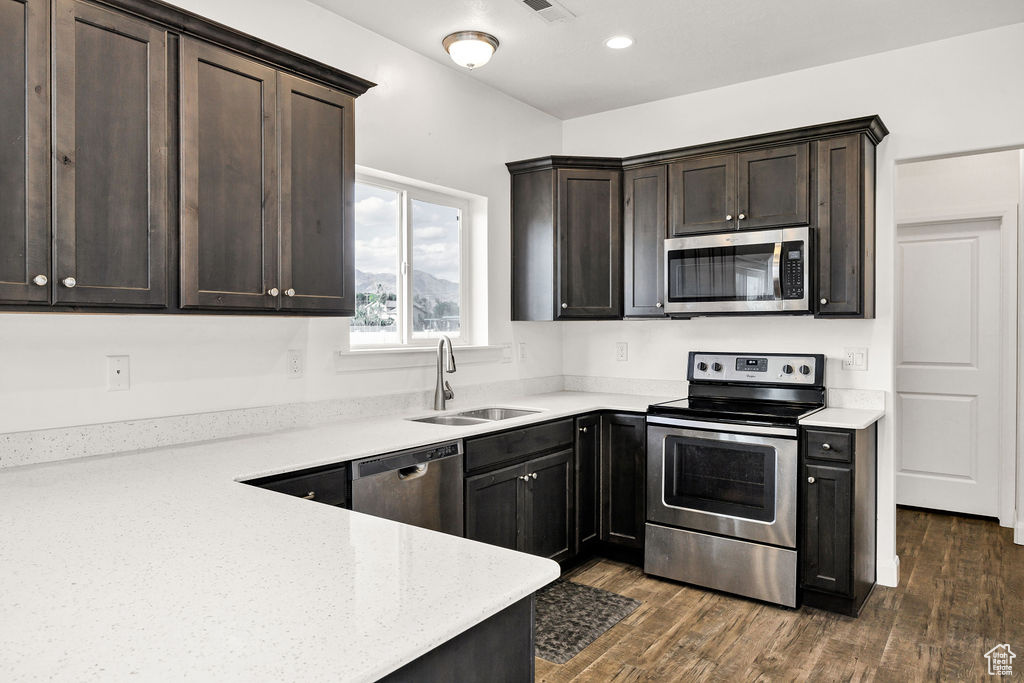 Kitchen with appliances with stainless steel finishes, a sink, dark brown cabinetry, dark wood-style floors, and light stone countertops