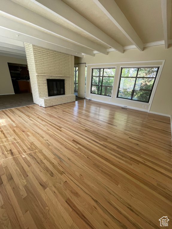 Unfurnished living room featuring beamed ceiling, light wood-type flooring, a brick fireplace, healthy amount of natural light, and baseboards