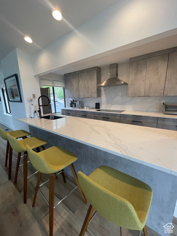 Kitchen with wall chimney range hood, a sink, a breakfast bar, black electric stovetop, and light stone counters