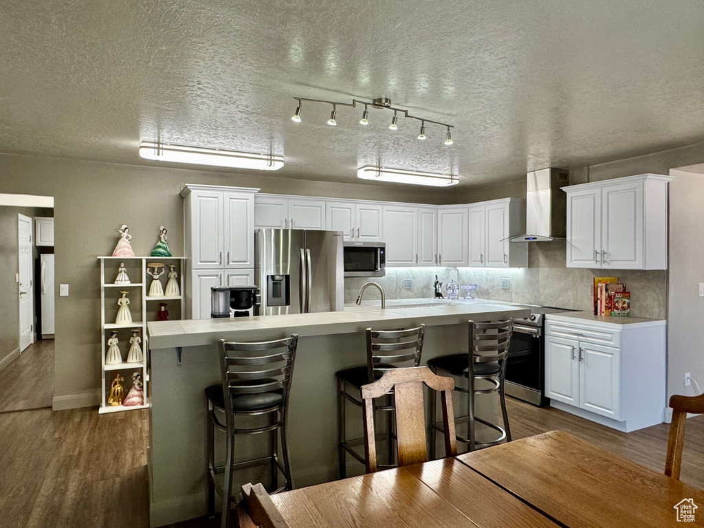 Kitchen featuring wall chimney range hood, stainless steel appliances, white cabinets, dark wood finished floors, and a textured ceiling