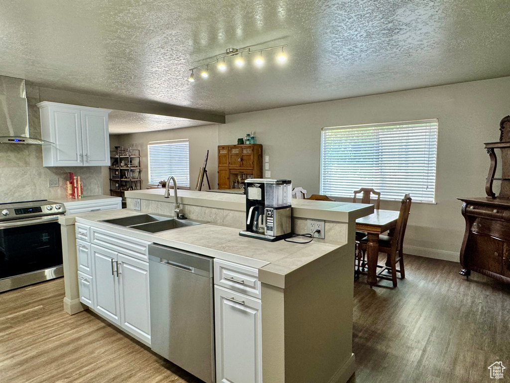 Kitchen with appliances with stainless steel finishes, a sink, wall chimney exhaust hood, light wood-style flooring, and a textured ceiling