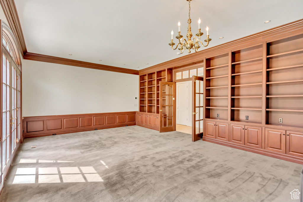 Carpeted empty room featuring crown molding, french doors, built in shelves, and an inviting chandelier