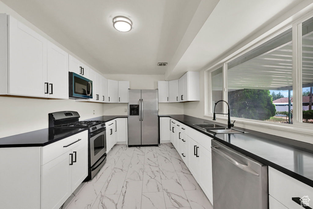 Kitchen featuring stainless steel appliances, a sink, light marble finish flooring, dark countertops, and white cabinetry