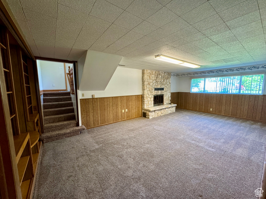 Unfurnished living room with wainscoting, wood walls, stairs, a stone fireplace, and carpet floors