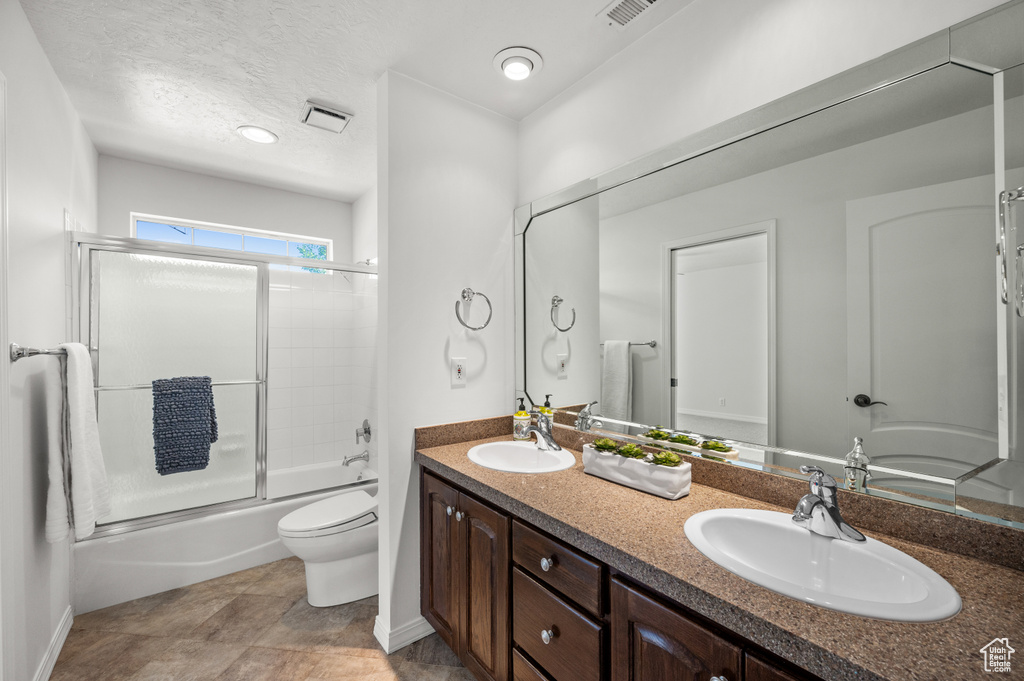 Full bathroom featuring toilet, double vanity, bath / shower combo with glass door, baseboards, and a textured ceiling