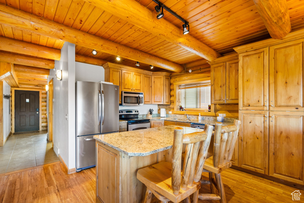 Kitchen featuring appliances with stainless steel finishes, a center island, wooden ceiling with exposed beams, light wood-style floors, and light stone counters