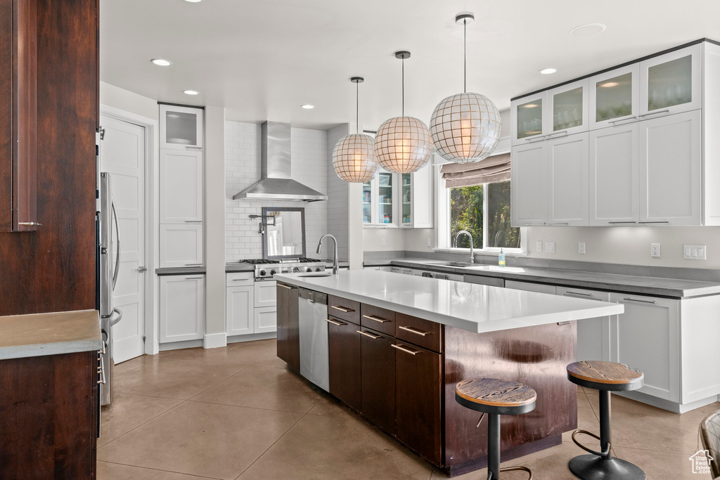 Kitchen featuring wall chimney exhaust hood, an island with sink, stainless steel dishwasher, tile patterned flooring, and glass insert cabinets