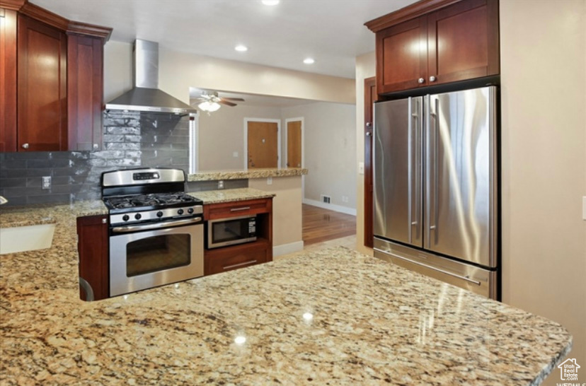 Kitchen with appliances with stainless steel finishes, wall chimney range hood, a peninsula, a ceiling fan, and backsplash