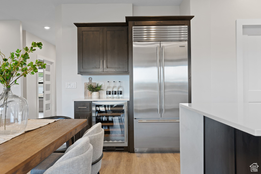 Kitchen with stainless steel built in fridge, beverage cooler, dark brown cabinetry, light countertops, and light wood-style flooring