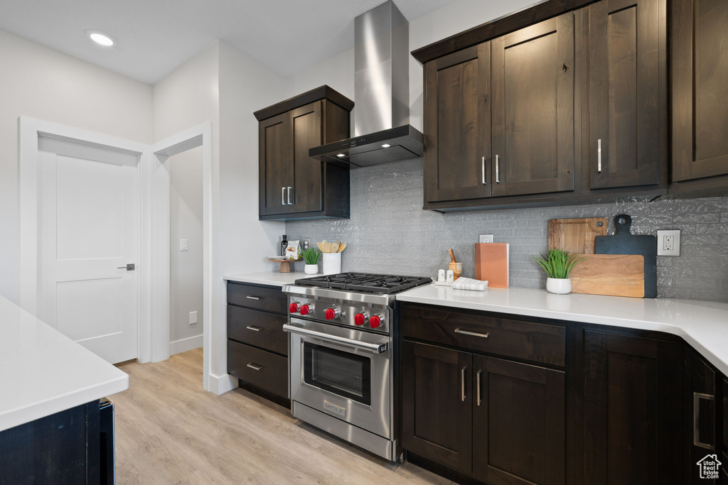 Kitchen with wall chimney range hood, luxury stove, decorative backsplash, dark brown cabinetry, and light wood finished floors
