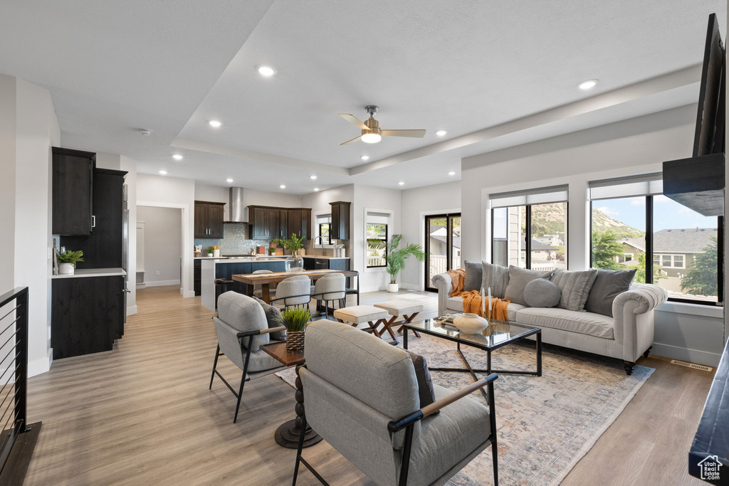 Living room with a raised ceiling, ceiling fan, light wood-style floors, baseboards, and recessed lighting
