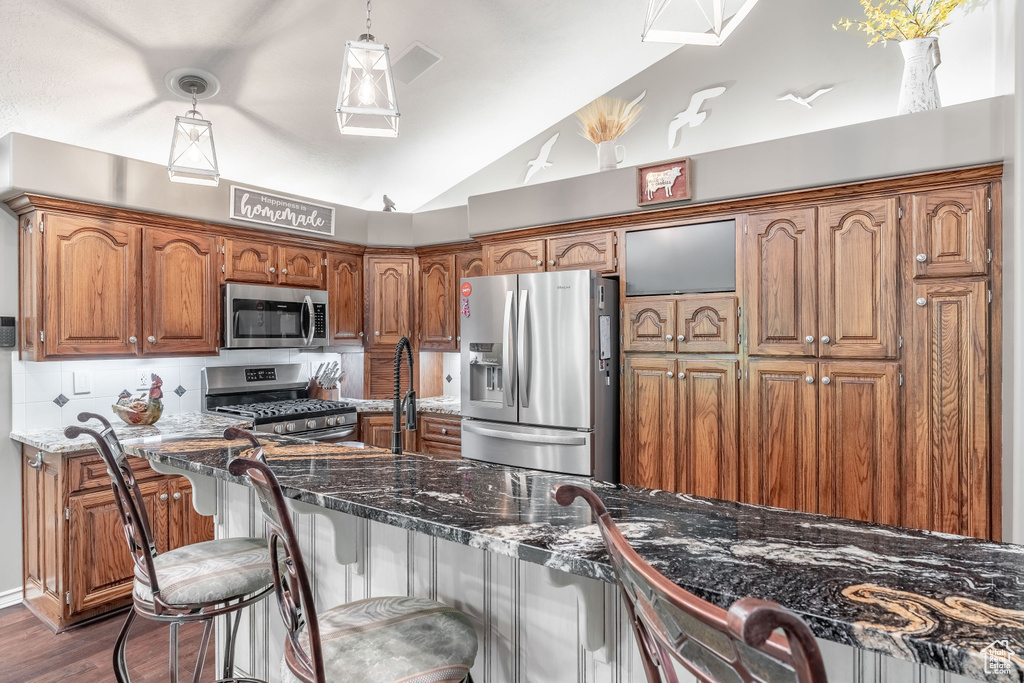 Kitchen featuring appliances with stainless steel finishes, lofted ceiling, brown cabinetry, dark wood-type flooring, and pendant lighting