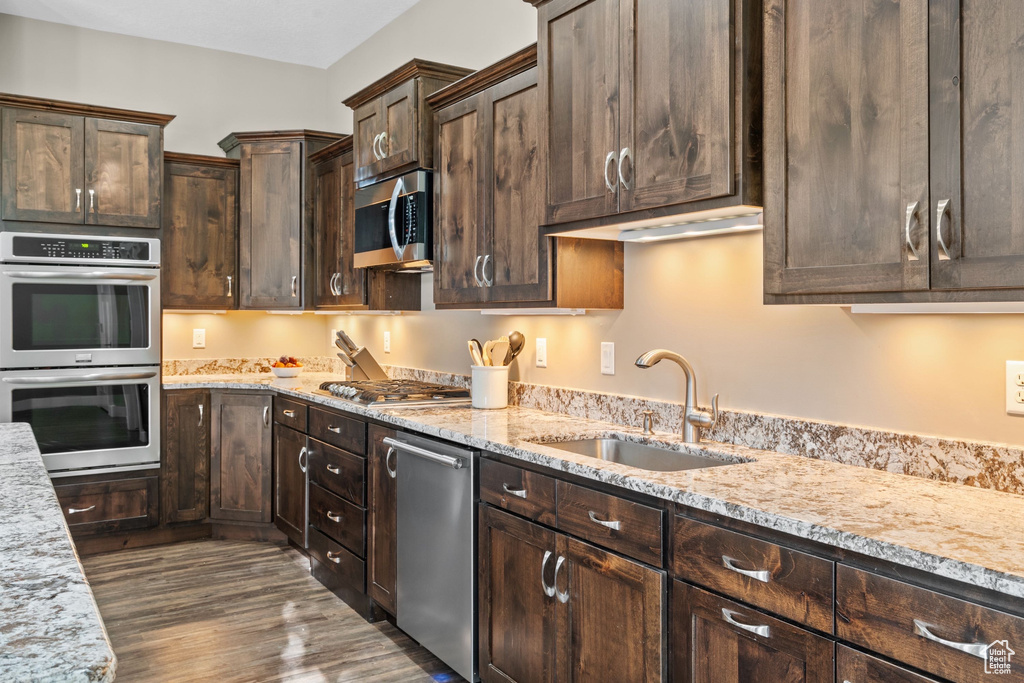 Kitchen featuring stainless steel appliances, a sink, dark wood-type flooring, dark brown cabinets, and light stone counters