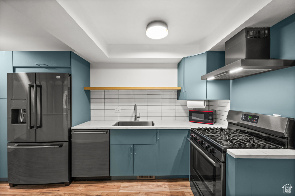 Kitchen featuring appliances with stainless steel finishes, a sink, wall chimney range hood, tasteful backsplash, and blue cabinets