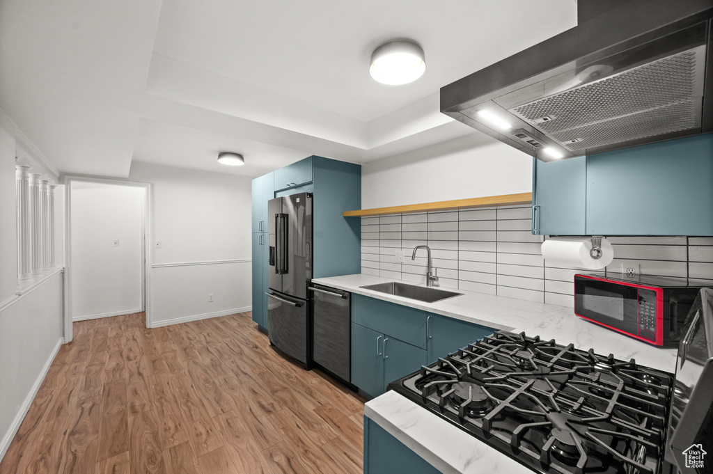 Kitchen with wall chimney exhaust hood, stainless steel appliances, blue cabinets, a sink, and light wood-style flooring