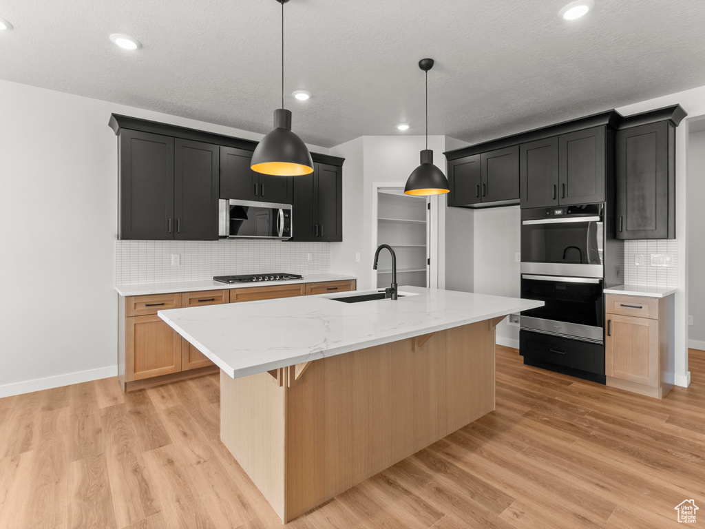 Kitchen with stainless steel appliances, decorative backsplash, light wood-type flooring, light stone counters, and a center island with sink