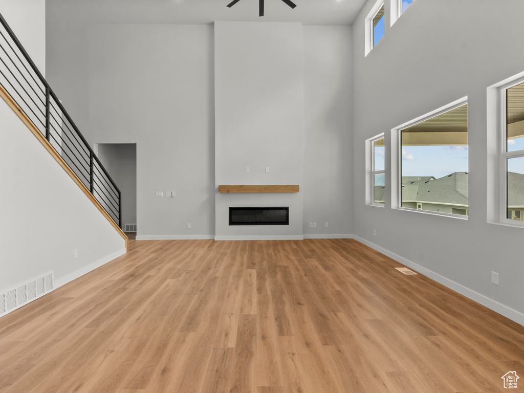 Unfurnished living room with a high ceiling, a ceiling fan, light wood-type flooring, a glass covered fireplace, and stairs