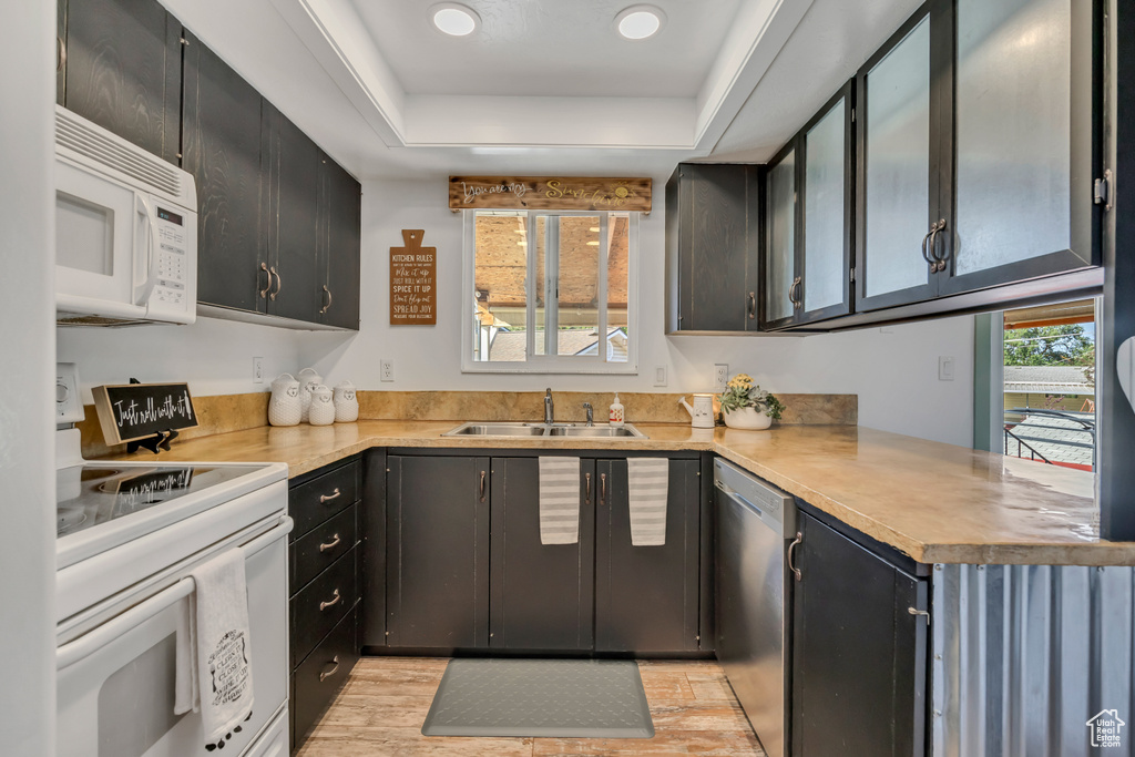 Kitchen with white appliances, a sink, a raised ceiling, dark cabinetry, and light countertops