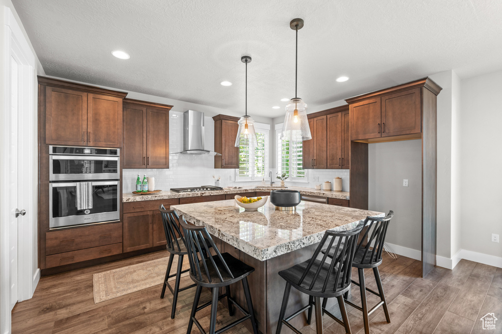 Kitchen featuring wall chimney range hood, appliances with stainless steel finishes, wood finished floors, decorative backsplash, and a kitchen island