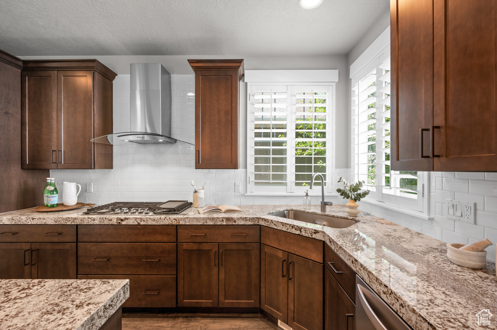 Kitchen with wall chimney range hood, stainless steel appliances, a sink, backsplash, and light stone countertops