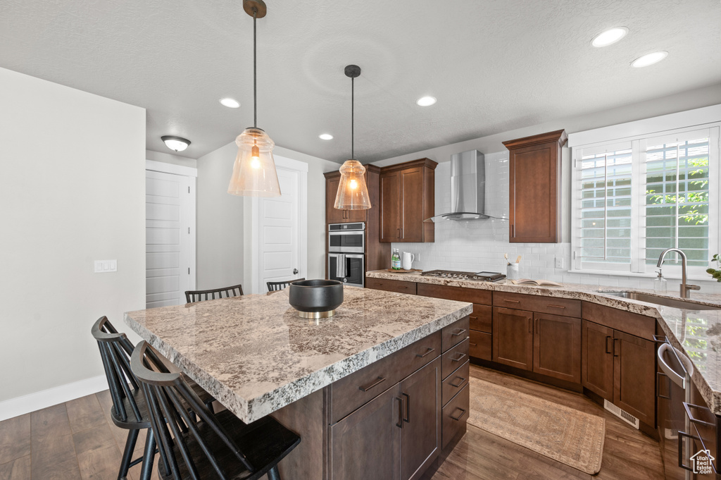 Kitchen with wall chimney range hood, a sink, appliances with stainless steel finishes, a kitchen bar, and dark wood-type flooring