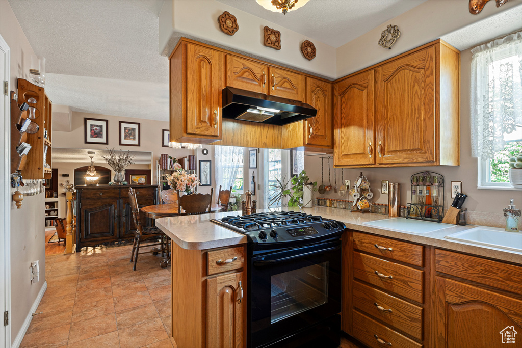 Kitchen with black range with gas cooktop, under cabinet range hood, a peninsula, brown cabinetry, and light countertops