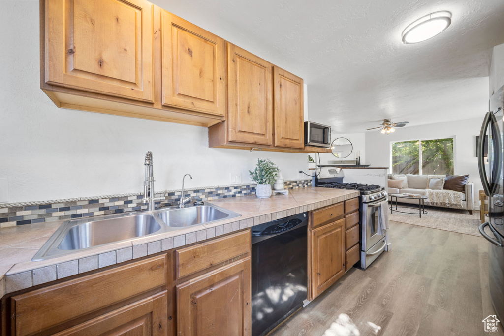 Kitchen featuring appliances with stainless steel finishes, a sink, tile counters, light wood-type flooring, and ceiling fan