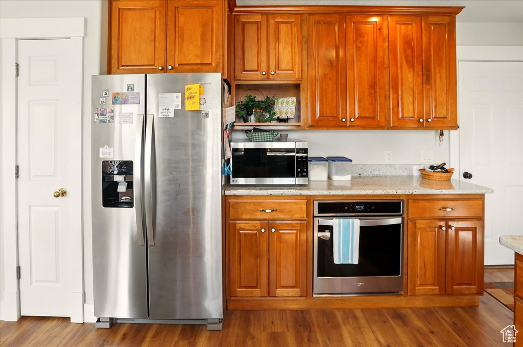 Kitchen featuring appliances with stainless steel finishes, wood finished floors, light stone counters, and brown cabinetry