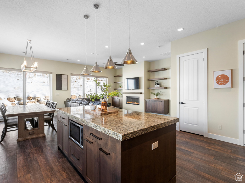 Kitchen featuring stainless steel microwave, dark wood-style flooring, dark brown cabinets, a glass covered fireplace, and decorative light fixtures