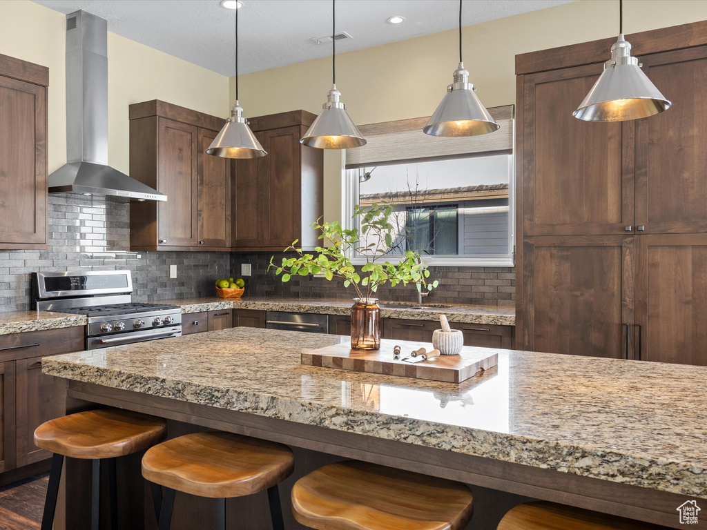 Kitchen featuring wall chimney range hood, stainless steel range with gas stovetop, a breakfast bar, tasteful backsplash, and light stone counters