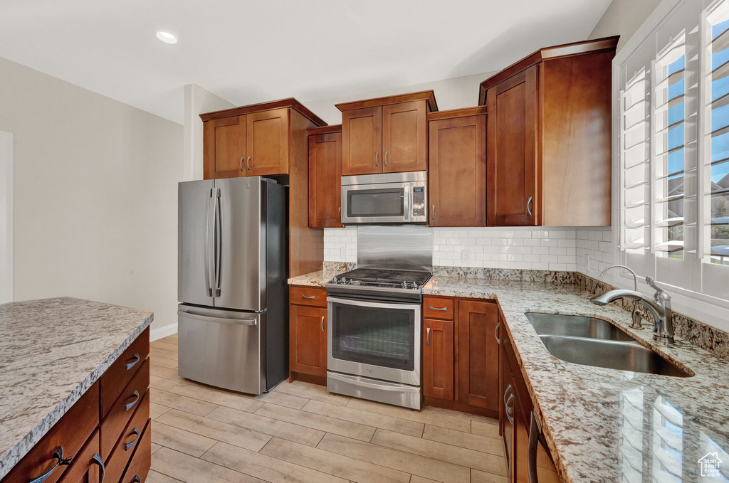 Kitchen with appliances with stainless steel finishes, a sink, backsplash, light stone counters, and brown cabinetry