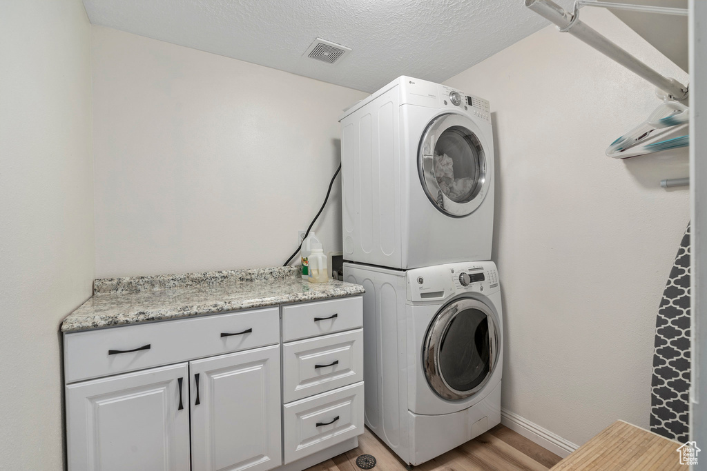 Laundry area with stacked washer / drying machine, cabinet space, light wood-style flooring, baseboards, and a textured ceiling