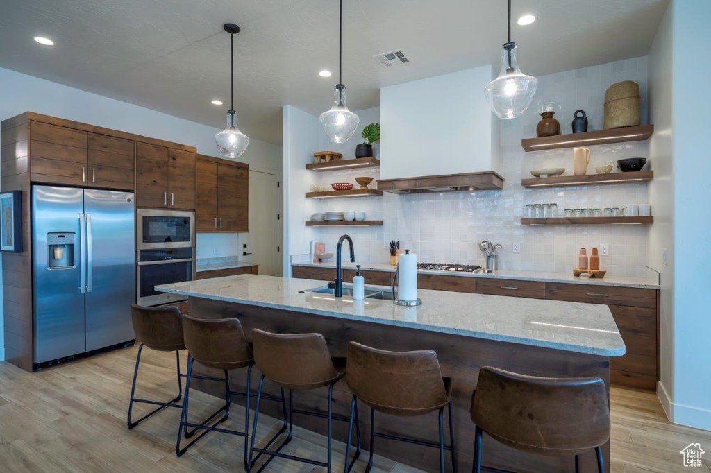 Kitchen featuring stainless steel appliances, open shelves, a sink, light wood-style flooring, and custom range hood