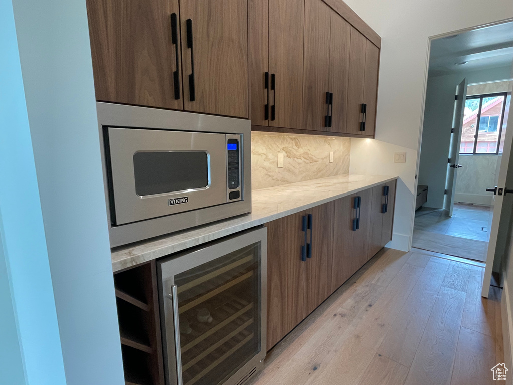 Kitchen with stainless steel microwave, beverage cooler, light wood-style flooring, tasteful backsplash, and light stone counters