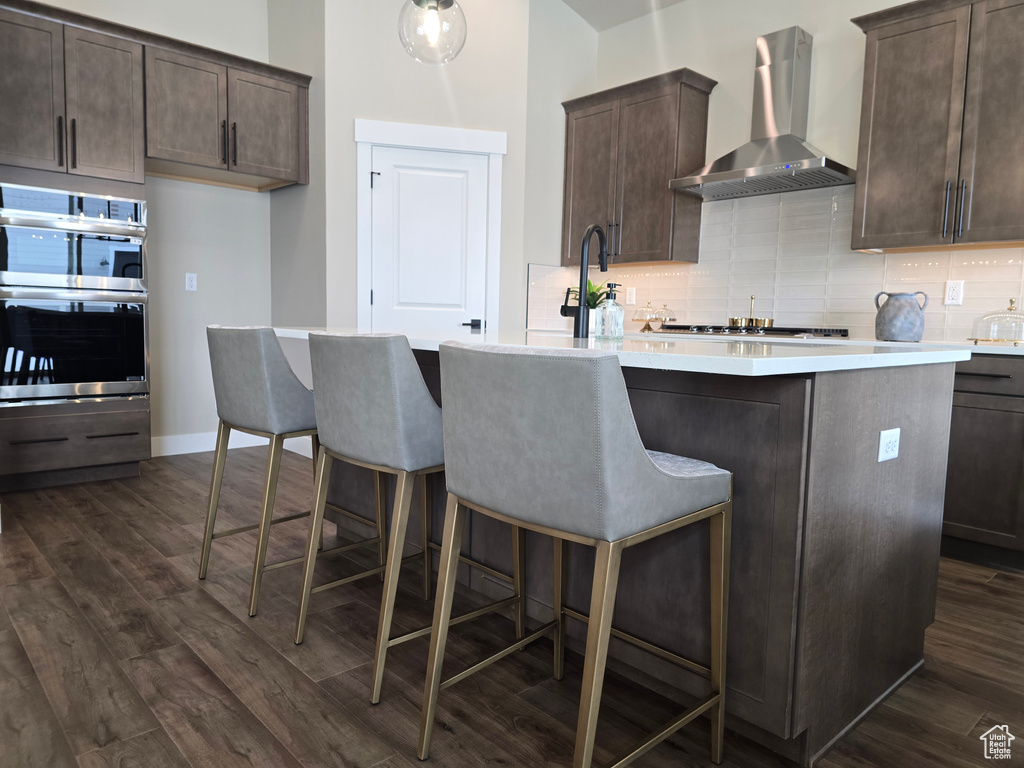 Kitchen featuring wall chimney range hood, stainless steel double oven, backsplash, and dark brown cabinets