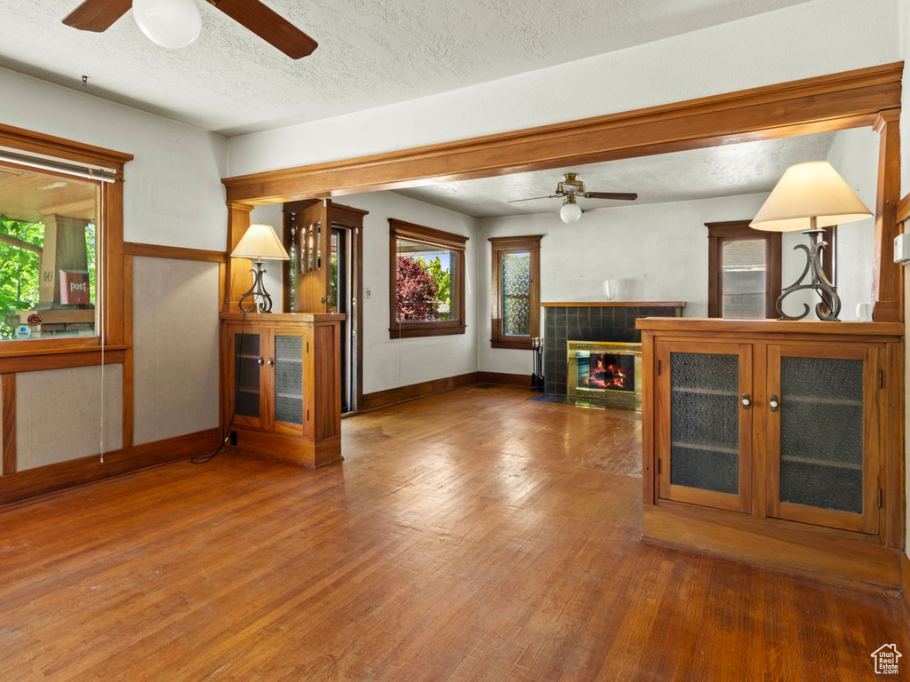 Unfurnished living room featuring a ceiling fan, wood finished floors, baseboards, a tiled fireplace, and a textured ceiling