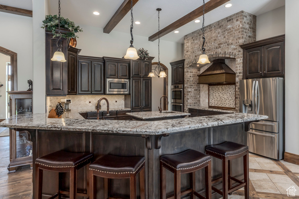 Kitchen with appliances with stainless steel finishes, beam ceiling, decorative backsplash, dark brown cabinets, and light stone countertops