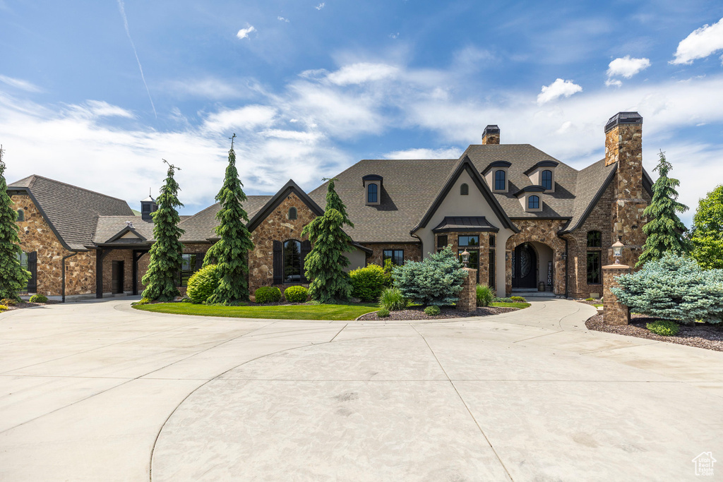 French country style house featuring curved driveway, stone siding, roof with shingles, and a chimney