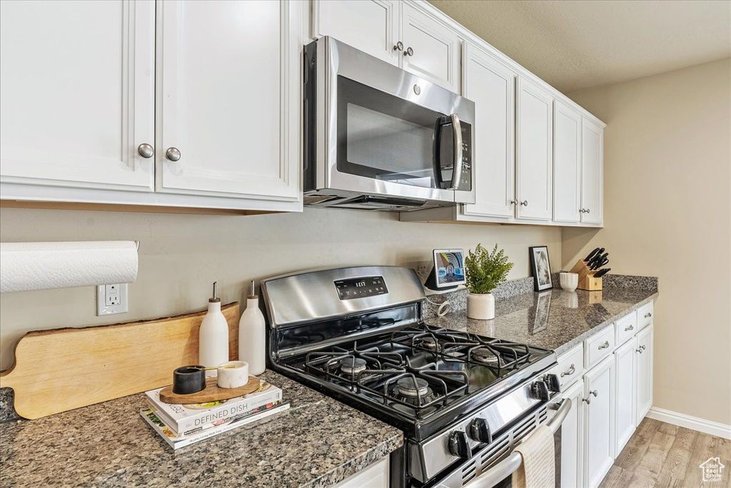 Kitchen with appliances with stainless steel finishes, white cabinetry, light wood-style flooring, dark stone countertops, and baseboards