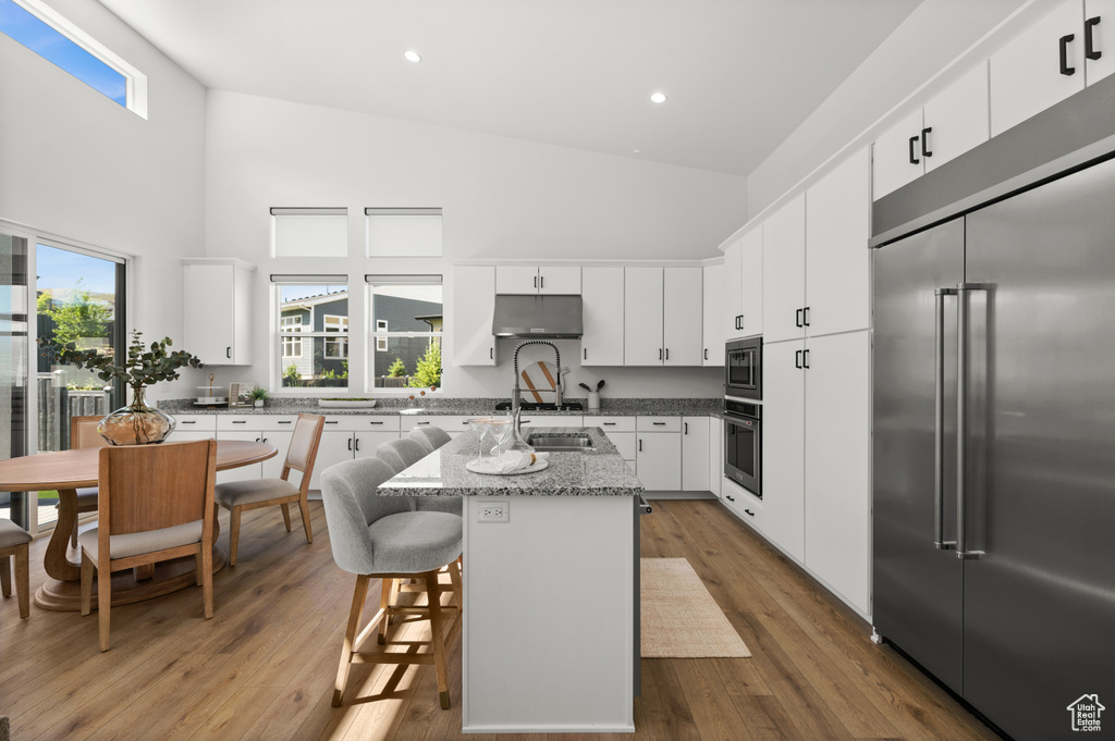 Kitchen featuring built in appliances, high vaulted ceiling, a kitchen island with sink, healthy amount of natural light, and white cabinets