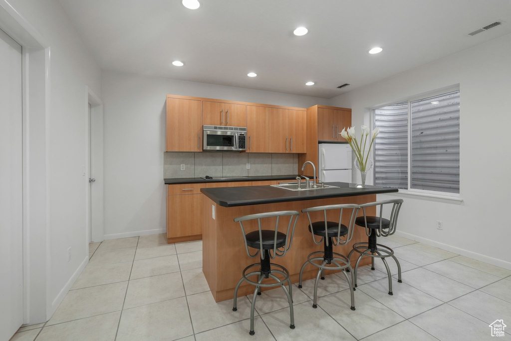 Kitchen featuring stainless steel microwave, a sink, dark countertops, an island with sink, and light tile patterned floors