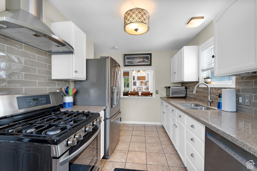Kitchen with stainless steel appliances, wall chimney range hood, a sink, light stone countertops, and white cabinetry