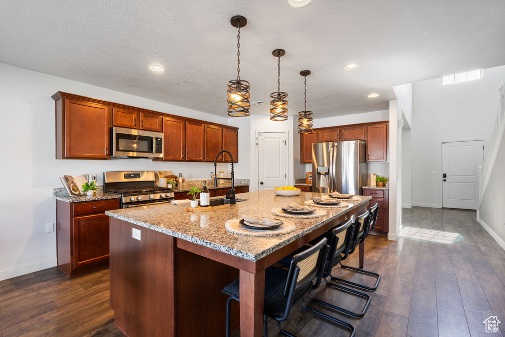 Kitchen featuring stainless steel appliances, an island with sink, light stone counters, dark wood-type flooring, and baseboards