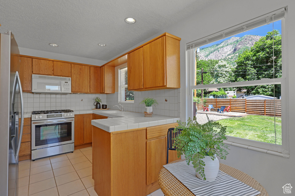 Kitchen with appliances with stainless steel finishes, backsplash, a peninsula, light tile patterned flooring, and healthy amount of natural light