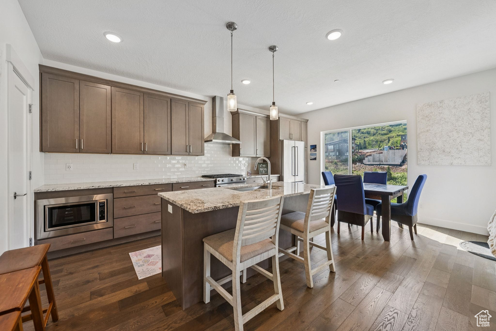 Kitchen featuring wall chimney range hood, stainless steel appliances, tasteful backsplash, a kitchen bar, and a kitchen island with sink
