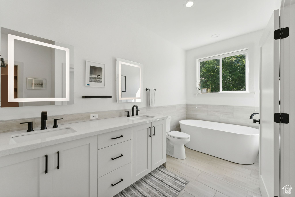 Full bathroom with double vanity, a freestanding tub, toilet, tile walls, and wainscoting